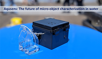 close up view of the Aqusens sensor, a small black box with a clear attachment, sitting on a blue table with the line "Aqusens: the future of micro-object characterization" across the top.