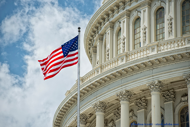 close-up view of U.S. Capitol dome, exterior, with an American flag waving ahead of it against a blue sky.